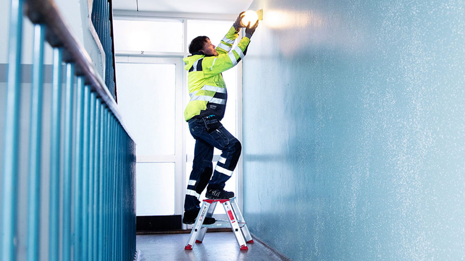 A janitor standing on a small step ladder to change the light bulb in a lamp.