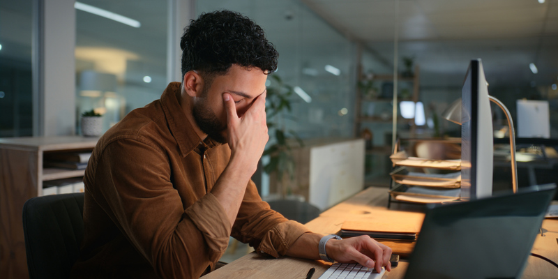 En yngre mand, der sidder ved sin computer på arbejde og tager sig til hovedet