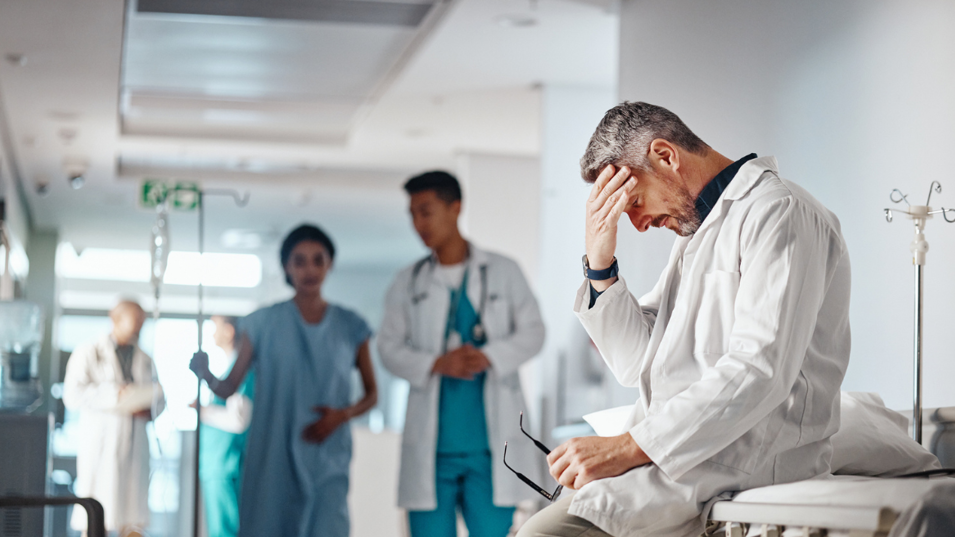 En læge der sidder på en hospitalsgang og tager sig til hovedet